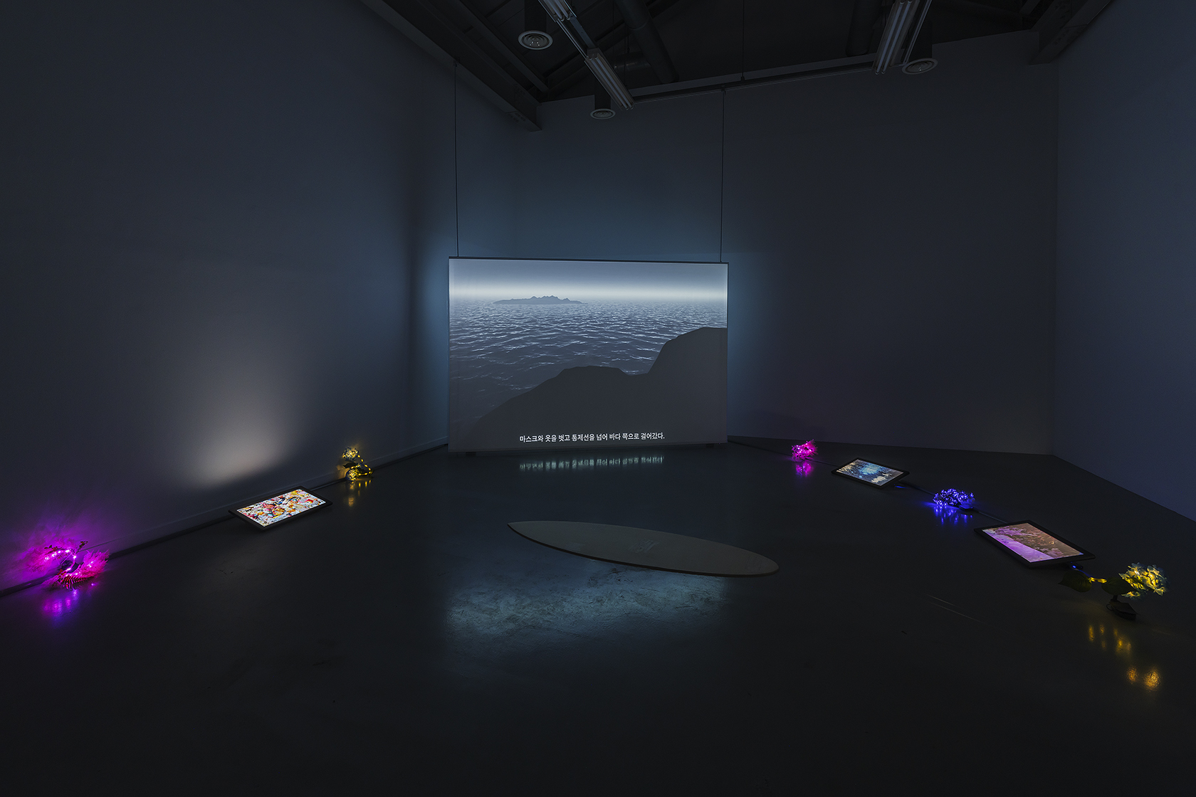 《退潮顯影》展場全景,錄像、衝浪板形瑜珈墊與發光人造花分布於暗室中。